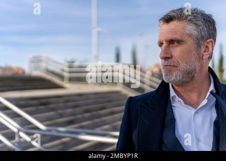 Confident bearded senior businessman in formal wear looking away while standing on stairs and thinking about project during walk Stock Photo