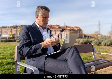 Concentrated senior businessman in formal wear checking notification on cellphone while surfing laptop during work on project on bench Stock Photo