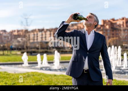Bearded middle aged businessman in formal suit drinking champagne from bottle while standing on street against fountain Stock Photo