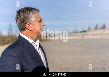 Side view of thoughtful senior businessman in formal clothes looking away while standing on street and admiring picturesque landscape Stock Photo