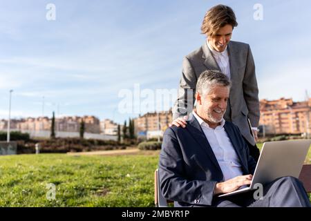 Positive businessmen in formal clothes smiling and working on remote project on laptop while spending time on street Stock Photo