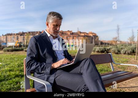 Focused mature businessman in formal suit working on project on laptop while sitting on bench on street Stock Photo