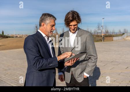 Smiling businessmen in formal suits browsing tablet while standing together on pavement and working on project Stock Photo