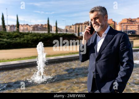Cheerful bearded mature businessman in formal wear smiling and looking away while talking on smartphone during walk near fountain Stock Photo