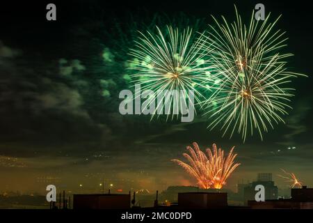 Colorful fireworks display over city at midnight. Green firework explosions and red fountains in the night sky while holiday celebration. Night scene. Stock Photo