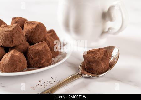 Chocolate truffle on a spoon closeup. Delicious dark chocolate candy balls in cocoa powder and white cup n a marble table. Sweet food confection. Stock Photo