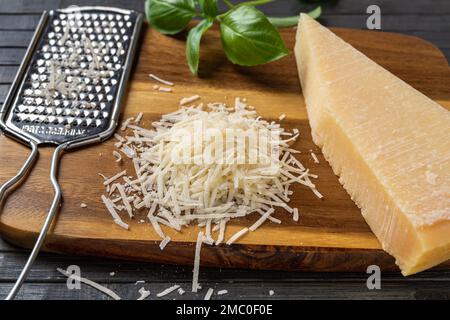 Heap of grated parmesan cheese closeup. Tasty grana padano cheese whole and grated and stainless steel grater on a wooden cutting board. Stock Photo