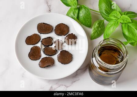 Sliced black truffle on a plate and jar of truffle carpaccio slices in oil over marble background. Canned delicacy mushroom Tuber melanosporum. Gourme Stock Photo