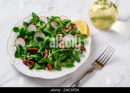 Lambs lettuce salad with radish and walnuts. Cornsalad leaves, nut kernels and sliced radish on white plate against marble surface. Low calories diet. Stock Photo