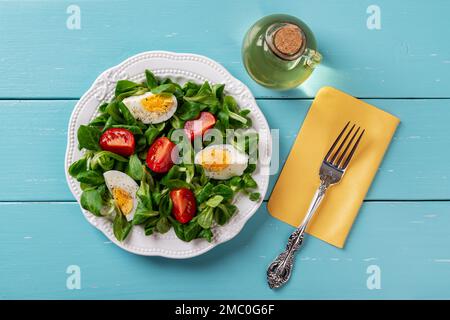 Salad of lambs lettuce, egg and tomato. Cornsalad leaves, quartered boiled egg and tomato on a white plate and fork over turquoise wooden table. Stock Photo