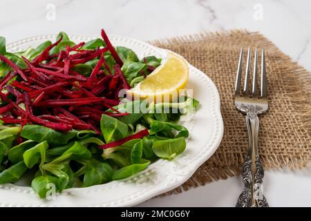 Slimming salad of lambs lettuce leaves and raw beet. Green corn salad and grated raw beet on a white plate and fork over marble surface. Vitamin salad Stock Photo