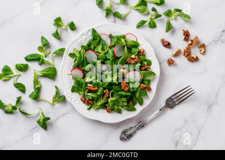 Salad of lambs lettuce, radish and walnuts. Cornsalad leaves, sliced radish and nut kernels on a white plate over marble surface. Slimming diet recipe Stock Photo