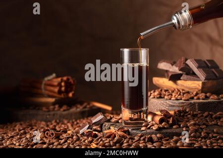 Coffee liquor is poured from a bottle into a glass. Coffee beans, cinnamon, anise, and pieces of bitter chocolate are scattered on the table. Stock Photo