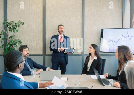caucasian leader with tablet in hand coaching interested business people, giving educational workshop presentation in Concentrated employees listening Stock Photo