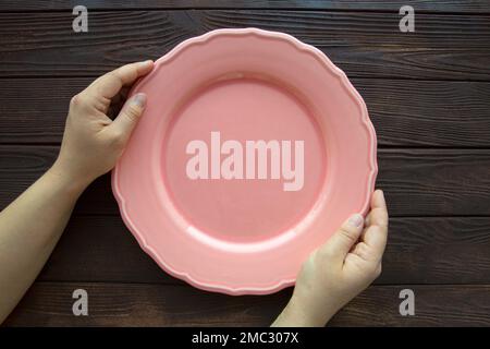 Empty pink plate hold by female hands, wooden background, top view. Diet or cooking concept. Stock Photo