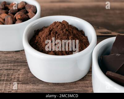 Cocoa powder in the bowl and other chocolate products on the wooden table, selective focus. Cacao powder, cocoa beans and dark chocolate Stock Photo