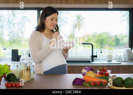 Beautiful Asian pregnant woman eating with lust cereals on breakfast in kitchen at home, enjoying meal. Yummy food for pregnant Stock Photo