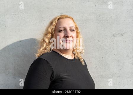 Portrait of a blonde and curly plus-size woman looking at camera while smiling over grey background Stock Photo