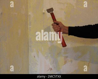 man's hand in black clothes holds a working tool with a narrow sharp metal tip and a long red handle against the background of a textured shabby room Stock Photo