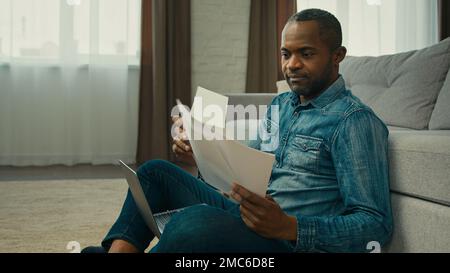 Mature african american man sitting in living room reading documents ethnic businessman freelancer working with papers analyzes finance checking Stock Photo