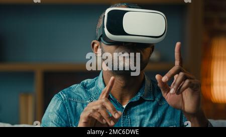 Close-up excited adult man in virtual reality glasses playing online game while sitting on couch in living room gesturing hands modern male gamer with Stock Photo