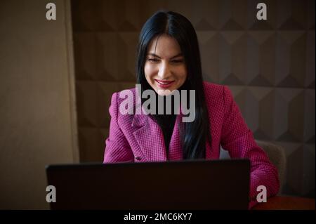 Smiling confident woman entrepreneur 40-45 years old, dressed in a stylish crimson jacket, talking to business clients via video link. Successful entr Stock Photo