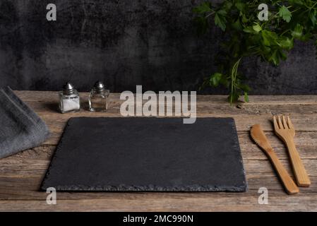 Empty black stone plate with wooden cutlery on a rustic old wooden table, next to salt, pepper and parsley leaves. Stock Photo