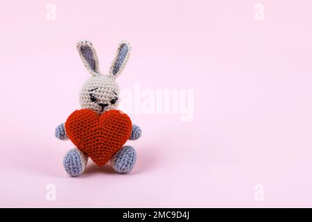 Knitted gray bunny with a crocheted red heart on a pink background. Happy Valentine's Day, Mother's Day and birthday greeting card. Stock Photo