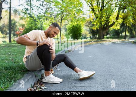 Man injured leg while jogging, african american man sitting on ground, massaging sore muscle with hands, upset and injured runner. Stock Photo