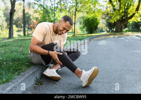 Man injured leg while jogging, african american man sitting on ground, massaging sore muscle with hands, upset and injured runner. Stock Photo