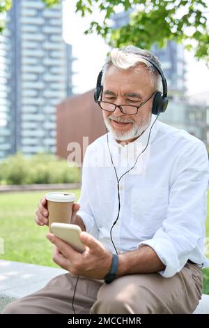 Older business man using smartphone wearing headphones having hybrid work call. Stock Photo