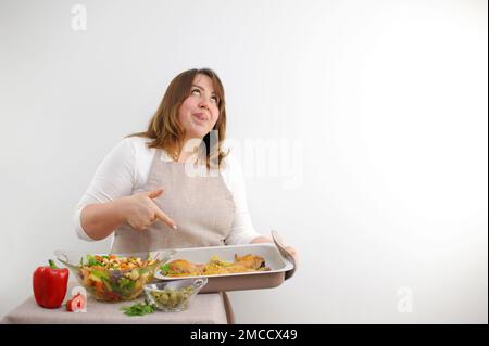 funny facial expression at cook puffy woman rolled eyes stuck tongue out licks lips with pleasure points to baking sheet in which dinner is cooked lunch brightly colorful salad with nuts ingredients Stock Photo