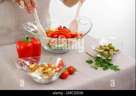 close-up of salad kneading with wooden spoons cherry tomatoes cocktail cucumbers olives and other ingredients lie on table with beige tablecloth beige apron restaurant food serving table setting Stock Photo