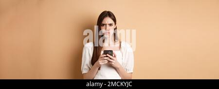 Disappointed girl with smartphone frowning, pucker lips upset, reading bad news on phone, standing on beige background Stock Photo
