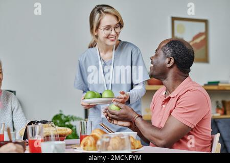 Smiling caregiver giving apples to senior man while he sitting at table and eating his breakfast Stock Photo