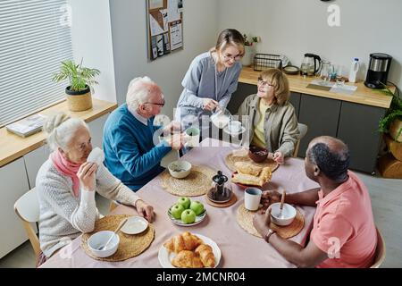 High angle view of young caregiver pouring milk in cups of senior people while they sitting at table and having breakfast Stock Photo