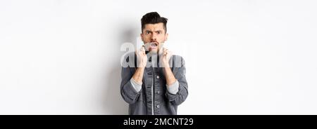 Shocked young man look confused and offended, gasping and frowning at camera, hear bad news, standing on white background Stock Photo