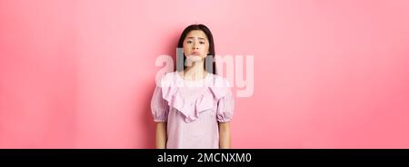 Sad whining asian girl pouting and frowning, looking upset about something unfair, complaining, standing in dress against pink background Stock Photo