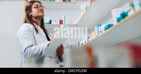 Female pharmacist writing on a clipboard while doing a stock take in a chemist. Woman standing in a drug store and counting inventory. Stock Photo
