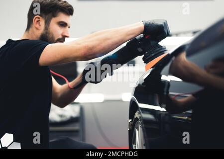 Man polishing black sports car paint varnish in car detailing studio. Side portrait. Blue collar worker. High quality photo Stock Photo