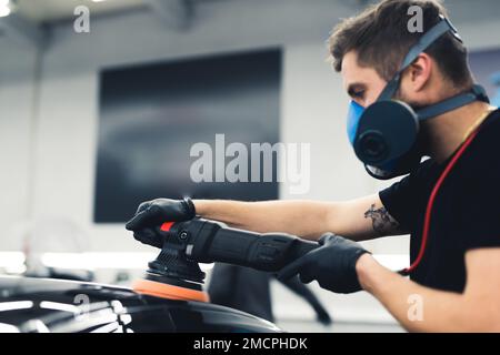 Man in protective mask polishing car paintwork with professional equipment - orbital polisher. Indoor medium closeup shot. High quality photo Stock Photo