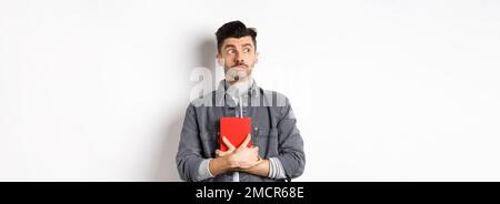 Silly young man looking aside at logo with timid face, holding his diary pressed to chest, carry journal, standing on white background Stock Photo