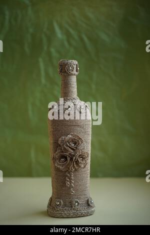 Decorative bottle with patterns and flowers made of ropes and threads in retro style on green old background. Vintage bottle. DIY. Souvenir toy on Stock Photo