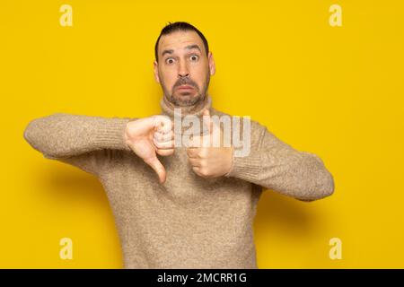 Bearded hispanic man wearing a turtleneck sweater giving a thumbs up and thumbs down in concept of doubt and indecision, isolated over yellow Stock Photo