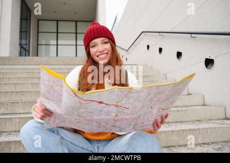 Young smiling redhead girl, tourist sits on stairs outdoors with city paper map, looking for direction, traveller backpacker explores city and looks Stock Photo