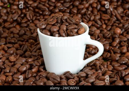 A white cup for espresso filled with whole coffee beans stands in a pile of roasted coffee beans Stock Photo