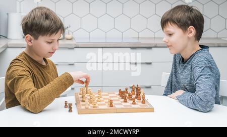 Side view of two calm focused preteen boys friends brothers sitting at white table in kitchen at home, playing chess on wooden board, competing. Board Stock Photo