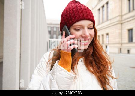 Mobile broadband and people. Smiling young redhead woman walks in town and talks on mobile phone, calling friend on smartphone, using internet to make Stock Photo