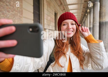 Cute young redhead woman takes selfie on street with mobile phone, makes a photo of herself with smartphone app on street Stock Photo