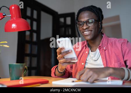 Happy optimistic African American man smm specialist with mobile phone sits at table in office Stock Photo